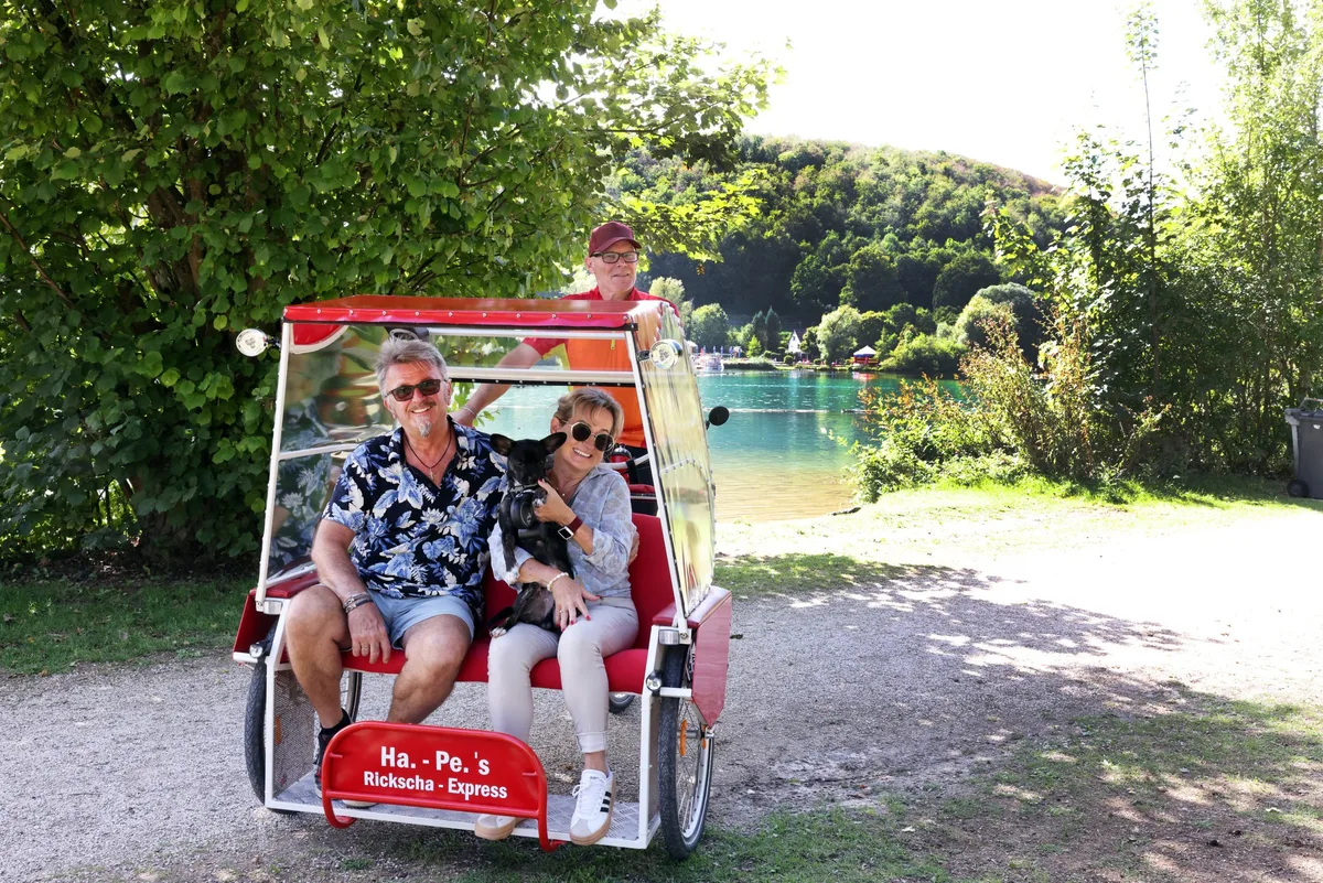Hans-Peter Brunners Rikscha-Express am Itzelberger See – Treffpunkt Imbiss auf der Friedhof-Seite Rikscha-Fahrer Hans-Peter Brunner mit Pia und Dietmar (Diddi) Pöche, den Pächtern des Imbisses am See, und ihrem Welpen Mailo.